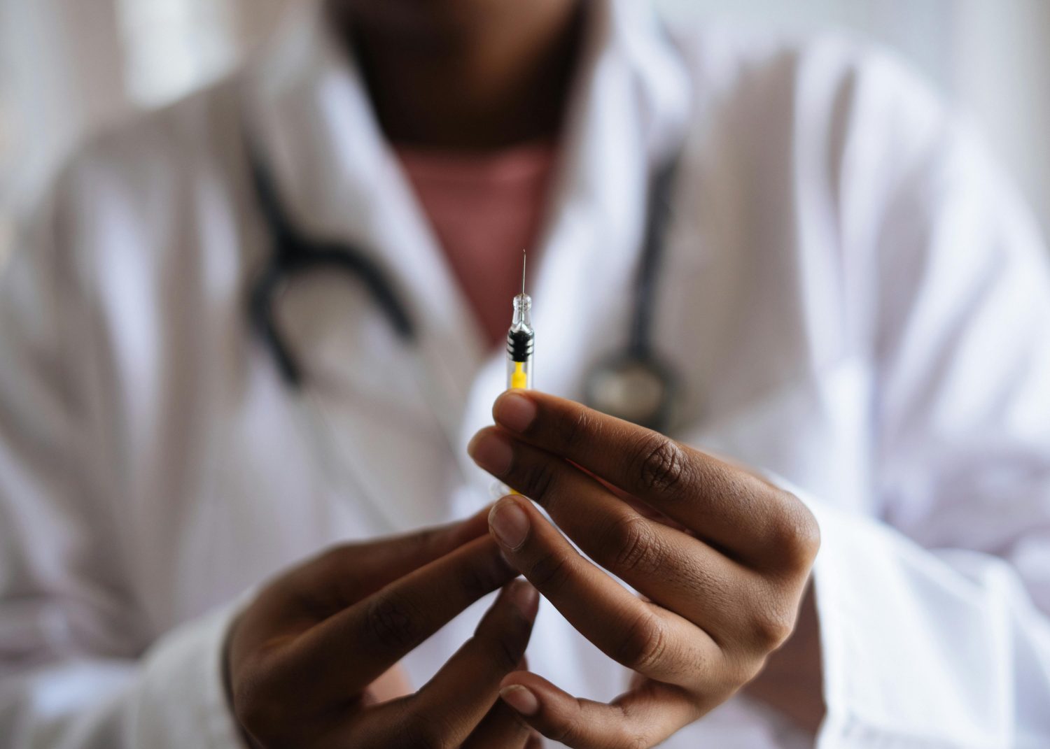 Eine Person in weißem Laborkittel und mit Stethoskop hält eine mit gelber Flüssigkeit gefüllte Spritze in der Hand und bereitet sich auf eine Injektion vor - möglicherweise Vitamin B12 zur Behandlung von Vitaminmangel. Der Fokus liegt auf der Spritze und den Händen, das Gesicht ist unscharf.