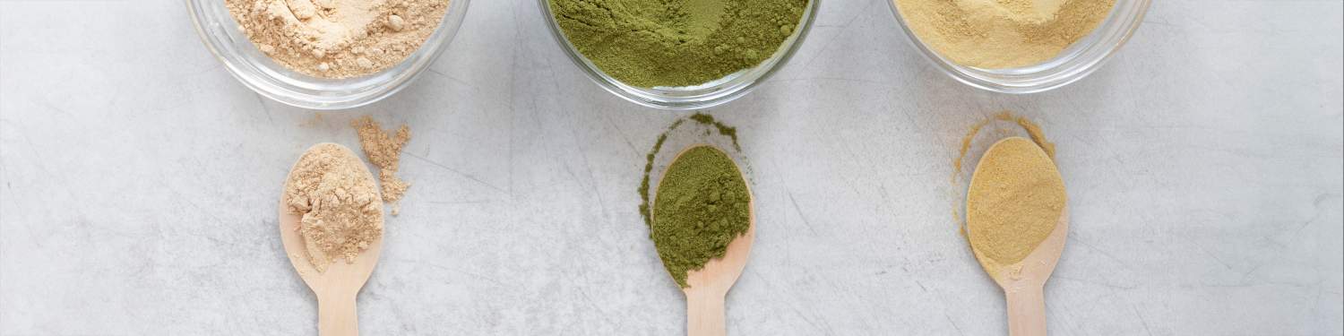 Drei Glasschalen gefüllt mit beigem, grünem und gelbem Pulver sind über drei Holzlöffeln mit dazu passenden Pulvern auf einer hellgrauen Oberfläche angeordnet und verdeutlichen so die Vielfalt der Produktion.