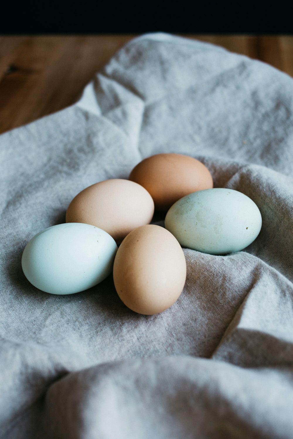 Fünf Eier in Hellbraun- und Hellblautönen sind auf einem weichen, beigen Tuch mit einer Holzoberfläche im Hintergrund angeordnet, inspiriert von Cholin Vida, unserem neuen Produkt für gesunde Ernährung.