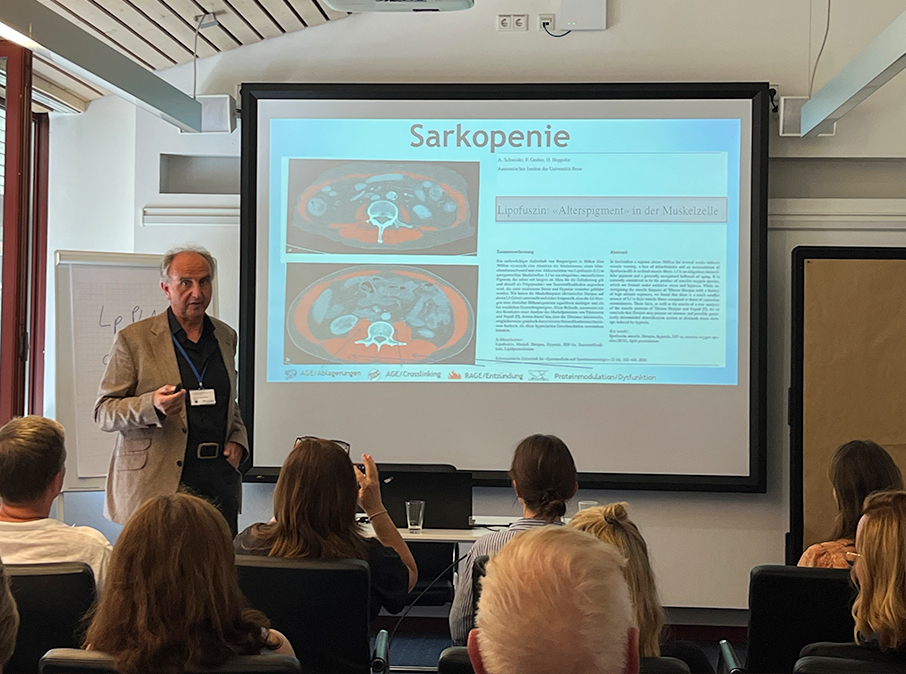 Ein Mann hält einen Vortrag über Sarkopenie und den AgeAway Shot vor einem Publikum, das in einem hell erleuchteten Konferenzraum neben einem Bildschirm steht, auf dem medizinische Bilder und Texte angezeigt werden.