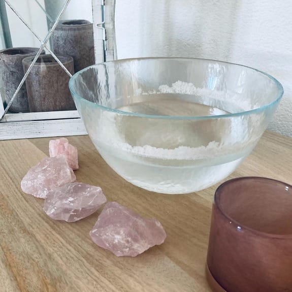 Eine mit Wasser gefüllte, klare Glasschale steht auf einer Holzoberfläche neben vier Rosenquarzkristallen und einer durchscheinenden violetten Tasse; ein Spiegel und zwei Kerzen im Hintergrund rufen die beruhigende Atmosphäre einer Naturheilpraxis mit Elementen von TEN hervor.