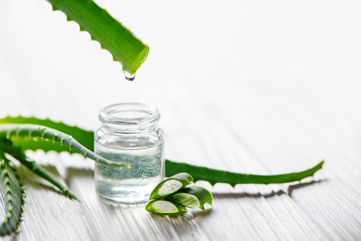 Ein kleines Glasgefäß, gefüllt mit Aloe-Vera-Gel, steht auf einer hellen Holzfläche. Einige aufgeschnittene Aloe-vera-Blätter umgeben das Glas, und ein Tropfen des Gels fällt von einem Blatt in das Glas.