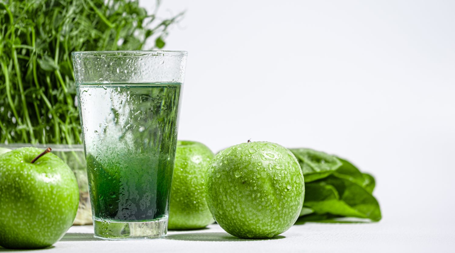 Ein Glas mit grünem Saft steht auf einer weißen Fläche, umgeben von frischen grünen Äpfeln, Blattspinat und grünen, chlorophyllreichen Kräutern. Auf den Äpfeln und dem Glas befinden sich Wassertröpfchen.
