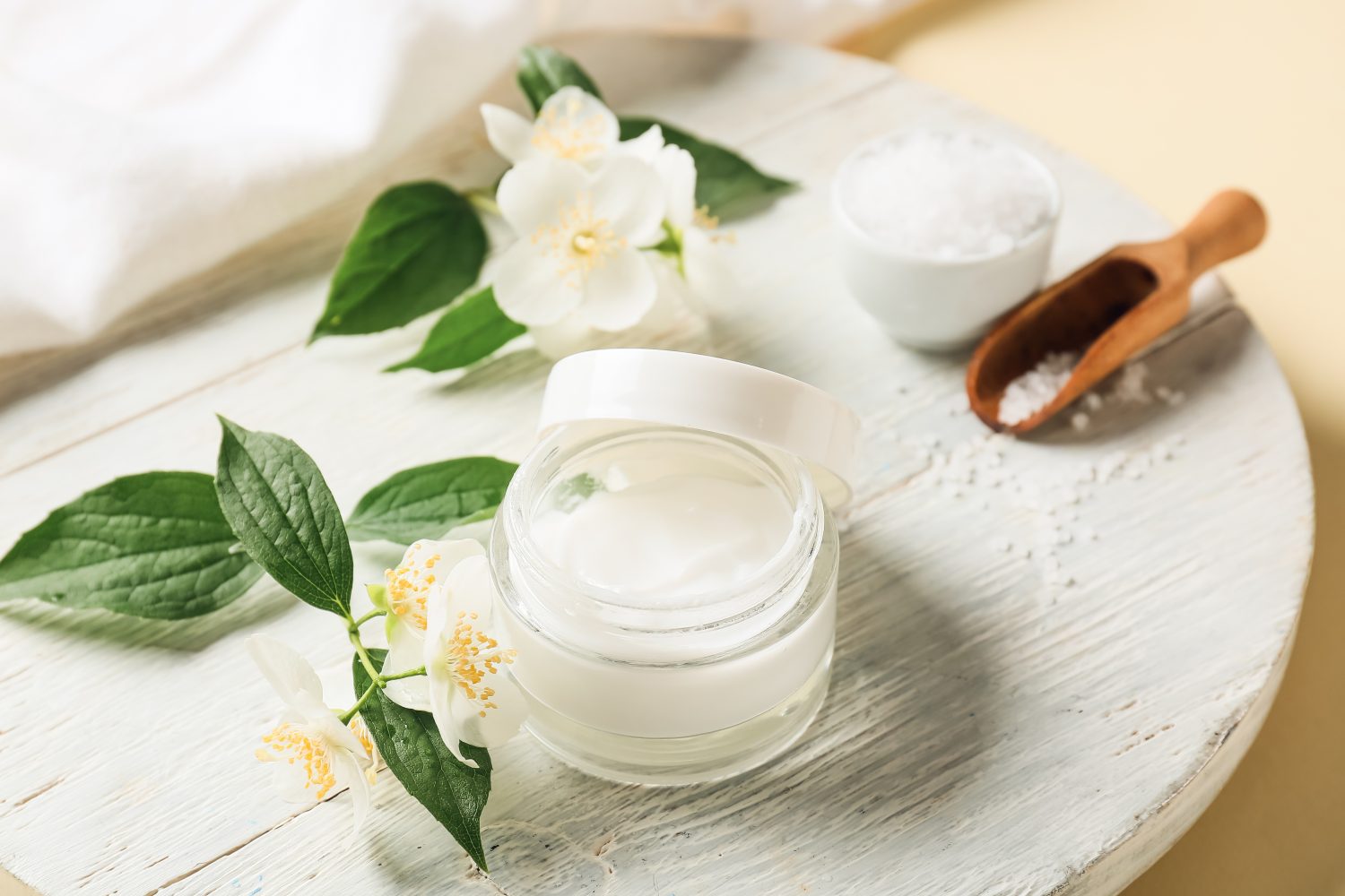 Geöffneter Tiegel mit weißer Naturkosmetik-Creme auf einem runden weißen Holztablett, dekoriert mit Jasminblüten, grünen Blättern, einer kleinen Schale mit Salz und einem Holzlöffel auf hellem Hintergrund.