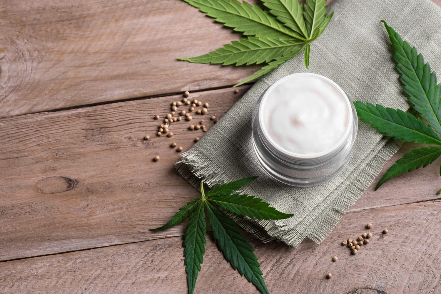 Ein Glas mit weißer CBD-Creme auf einem grünen Tuch, umgeben von Cannabisblättern und -samen, die auf einer Holzunterlage stehen.