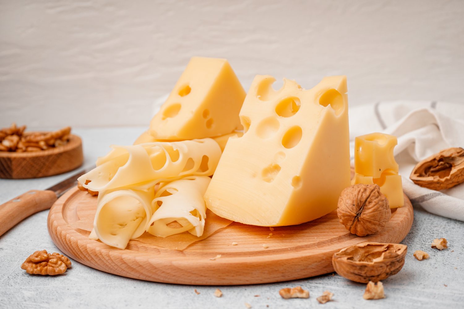 Mehrere Stücke Schweizer Käse mit großen Löchern, teils in Scheiben, teils im Ganzen, sind auf einem Holzbrett angeordnet, auf dem Walnüsse verstreut sind. Ein weißes Tuch und weitere Walnüsse befinden sich im Hintergrund.