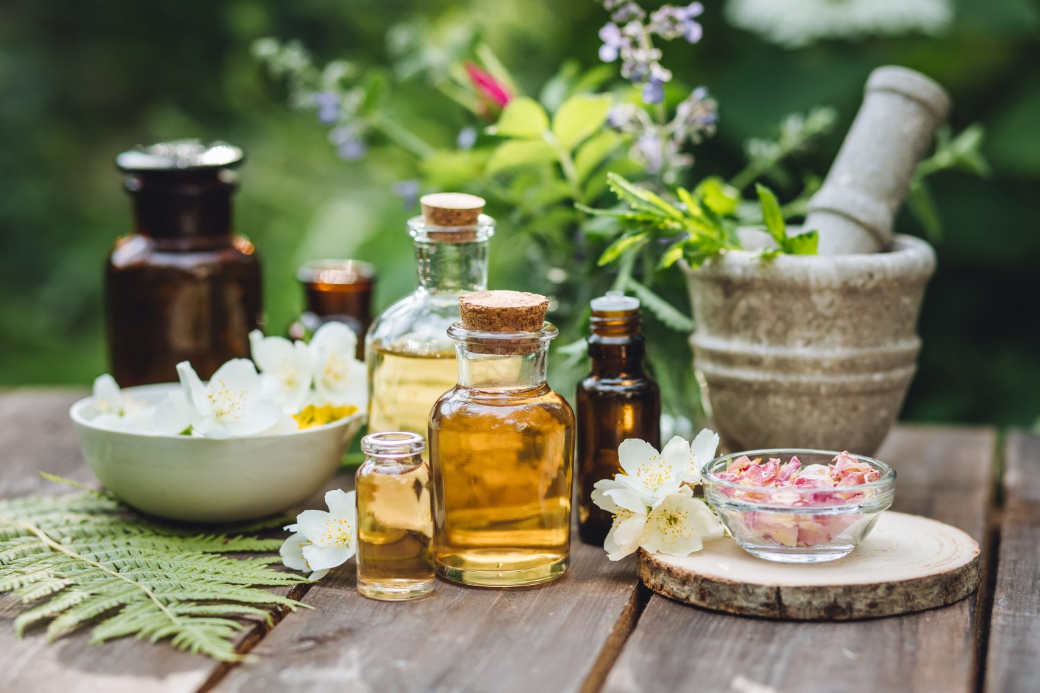 Eine Sammlung von Glasflaschen mit Kräuterölen, Naturkosmetik-Essentials, einer Schale mit Blütenblättern, frischen Blumen und einem Mörser und Stößel aus Stein, arrangiert auf einem Holztisch im Freien mit Grünzeug im Hintergrund.