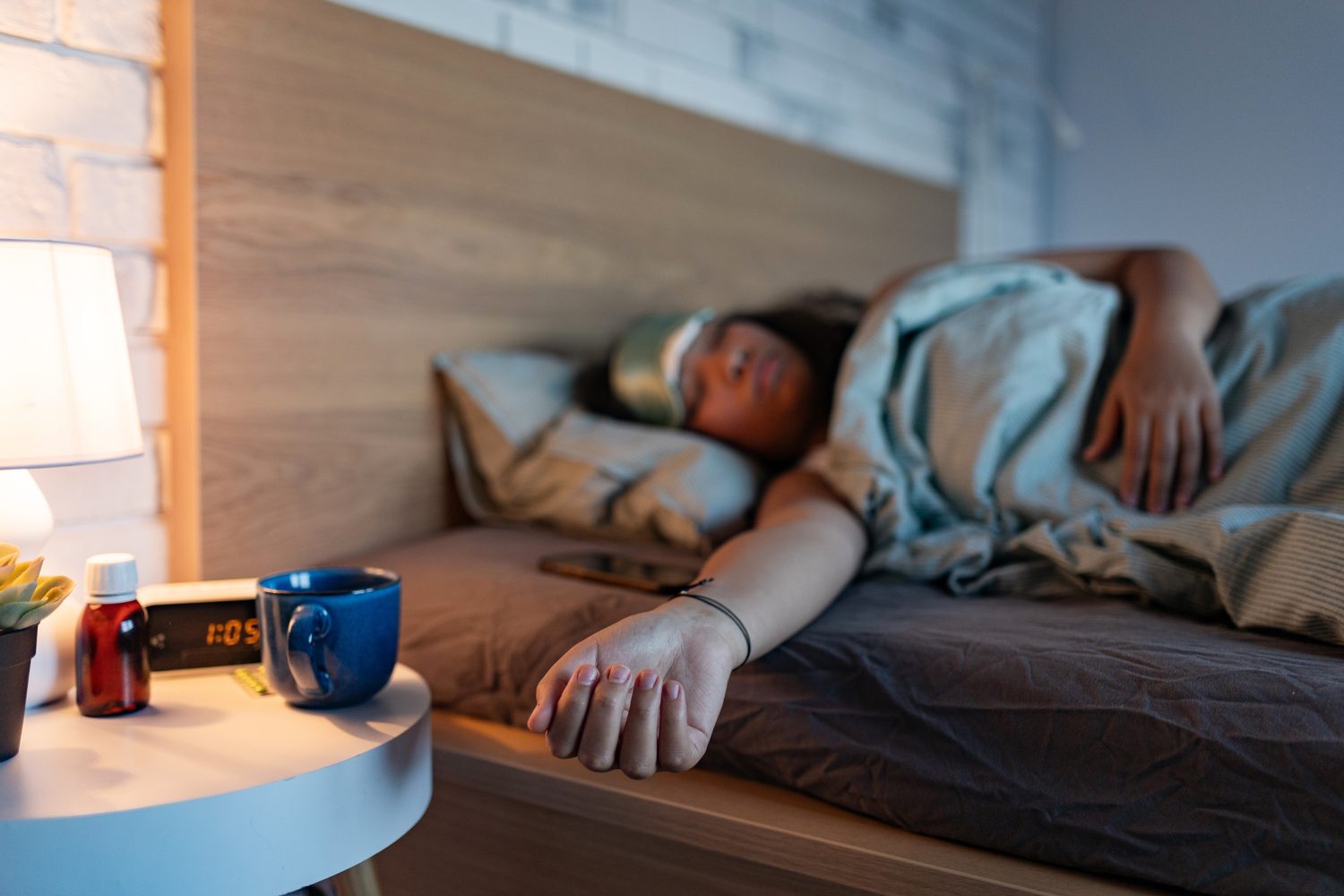 Eine Person schläft im Bett, ein Arm hängt über die Seite, sie trägt eine Augenmaske. Auf dem Nachttisch stehen eine Lampe, ein blauer Becher, Medizinflaschen und eine Uhr, die 11:05 Uhr anzeigt. Das Zimmer ist schwach beleuchtet, was eine gemütliche Atmosphäre schafft.