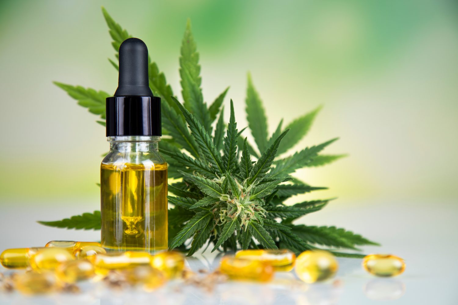 Eine kleine Glastropfflasche mit CBD-Öl steht neben einem frischen Cannabisblatt und mehreren gelben Kapseln, die alle auf einer spiegelnden Oberfläche mit einem grünen, verschwommenen Hintergrund angeordnet sind, der die beruhigenden Eigenschaften von CBD hervorhebt.
