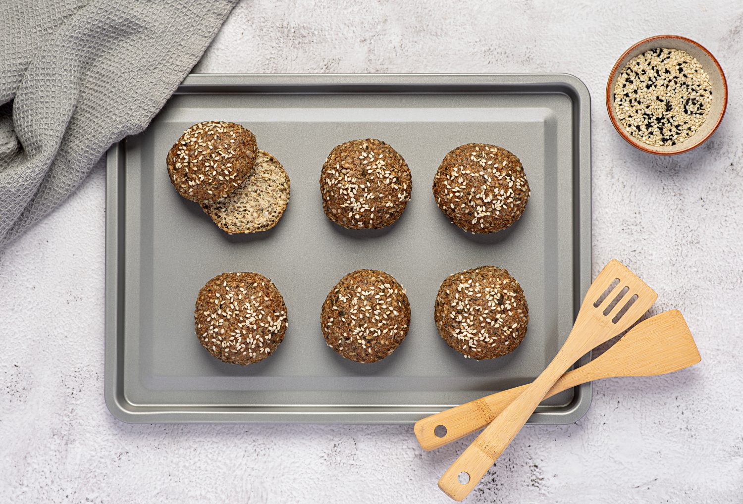 Sechs runde, braune, mit Sesam, Mohn und Flohsamenschalen belegte Brötchen sind auf einem Backblech angeordnet. Zwei hölzerne Küchenutensilien und eine Schale mit gemischten Samen stehen daneben, in der Ecke liegt ein graues Tuch.