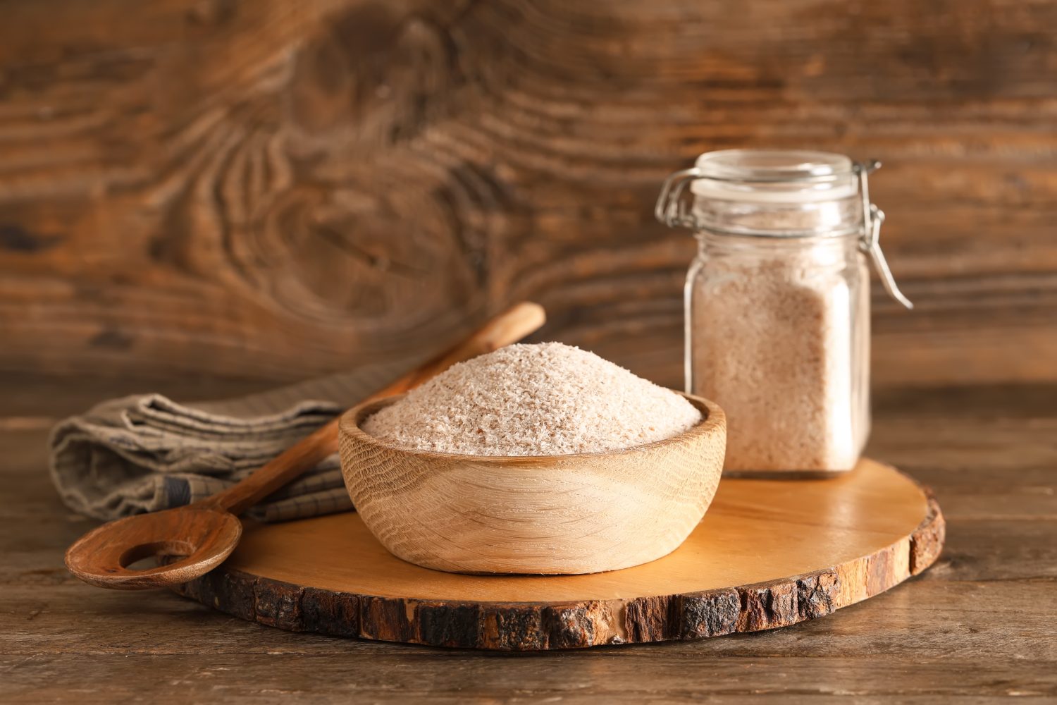 Eine mit rosa Himalaya-Salz gefüllte Holzschale steht auf einem Holztablett, daneben ein Glas mit demselben Salz, ein Holzlöffel, Flohsamenschalen und ein gefaltetes Tuch, alles auf einer rustikalen Holzfläche.