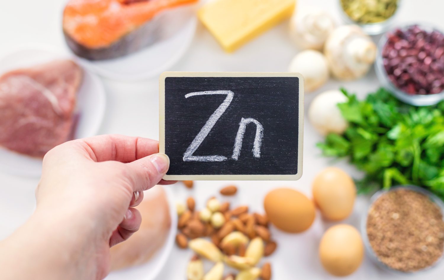 Eine Hand hält eine kleine Kreidetafel mit der Aufschrift Zn, umgeben von zinkhaltigen Lebensmitteln wie Fleisch, Fisch, Eiern, Käse, Nüssen, Samen, Pilzen und Blattgemüse auf einer weißen Fläche.