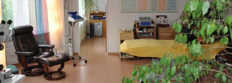 Ein heller Praxis- oder Therapieraum mit einem gelb bezogenen Untersuchungstisch, einem schwarzen Ruhesessel, medizinischen Geräten, einer Topfpflanze und bunten Vorhängen am Fenster. Schautafeln und Regale schmücken die Wände und schaffen eine einladende Naturheilkunde-Atmosphäre.