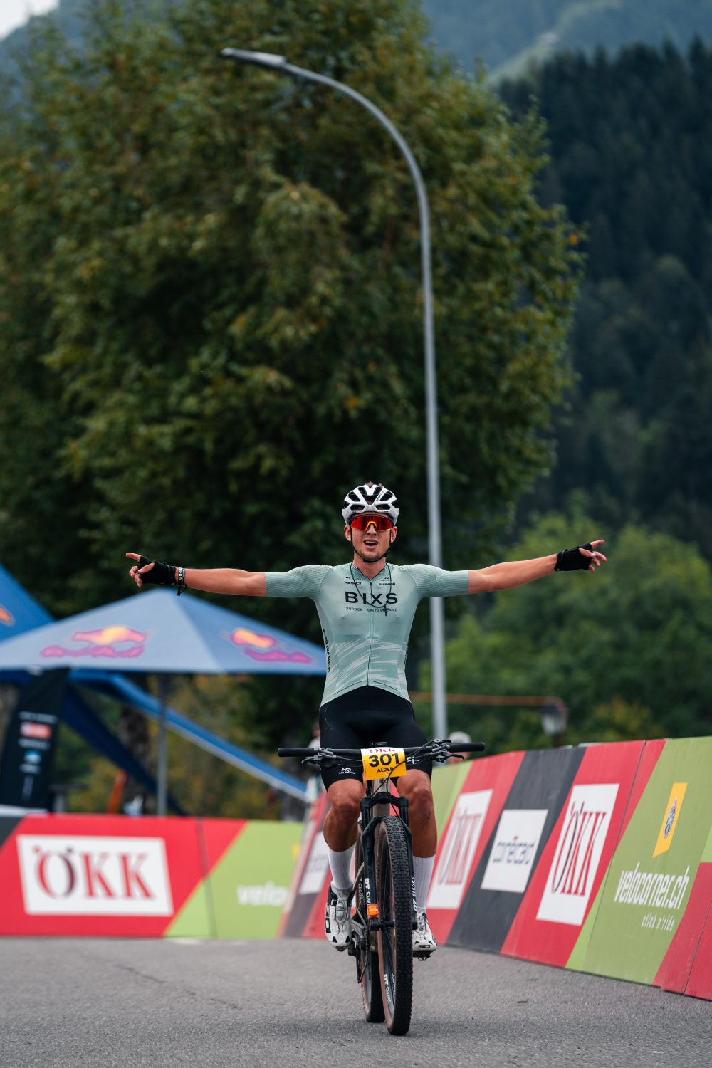 Ein Radfahrer mit Helm und grünem Trikot überquert die Ziellinie mit zum Sieg erhobenen Armen. Im Hintergrund sind Bäume, Berge und Veranstaltungsbanner am Straßenrand zu sehen.