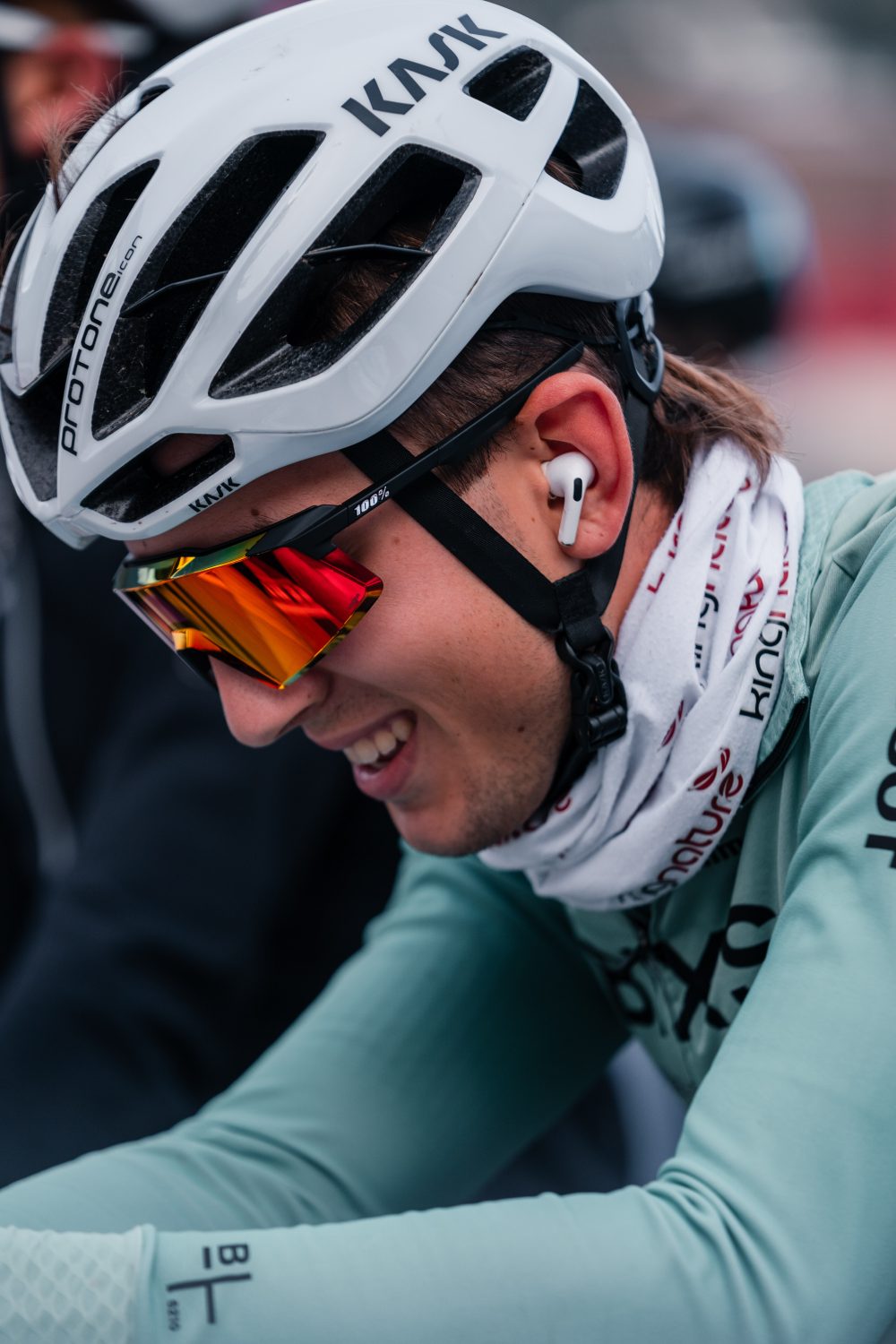 Ein Radfahrer, der einen weißen Helm, eine Sonnenbrille und eine weiße Halskrause trägt, lächelt und lehnt sich nach vorne. Er ist mit einem hellgrünen, langärmeligen Trikot bekleidet und hat drahtlose Ohrhörer in den Ohren.