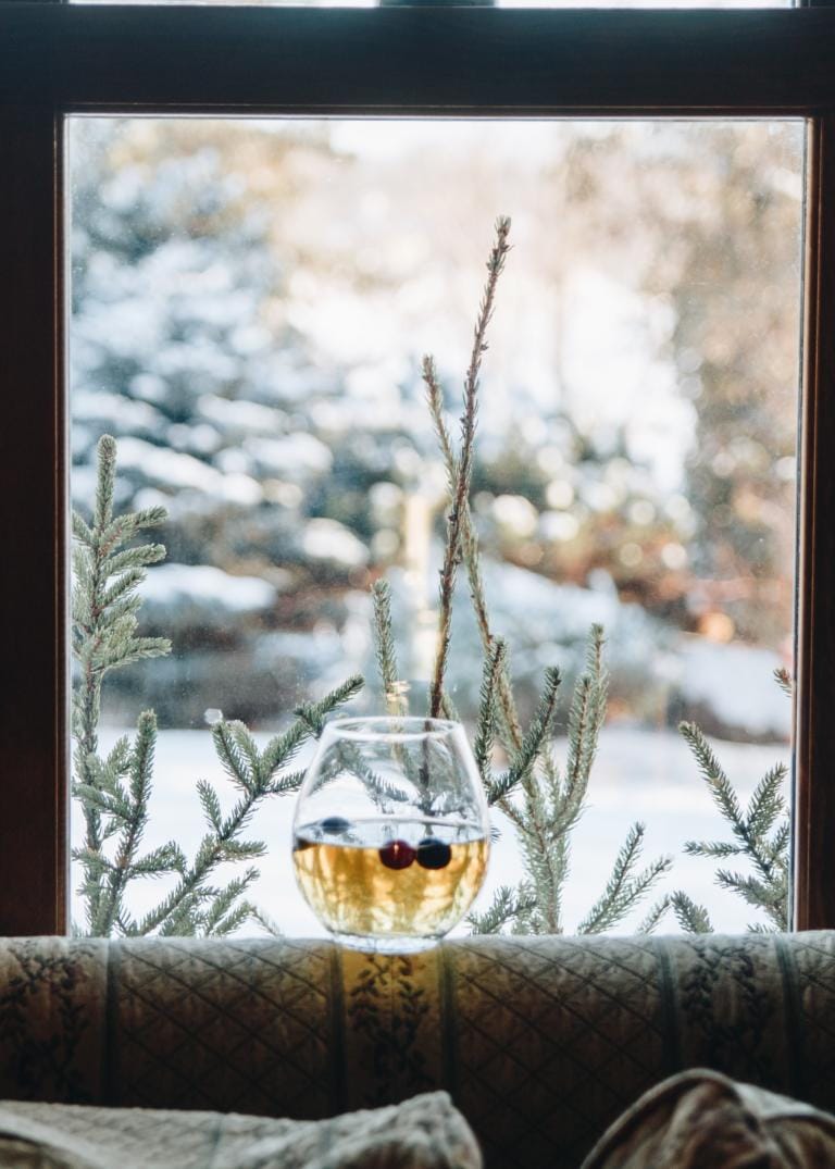 Ein Glas eines hellen Getränks mit Beeren steht auf einer Fensterbank vor einem schneebedeckten immergrünen Baum und einem bereiften Fenster und fängt eine gemütliche Winterlandschaft mit erfrischenden Aromen ein.