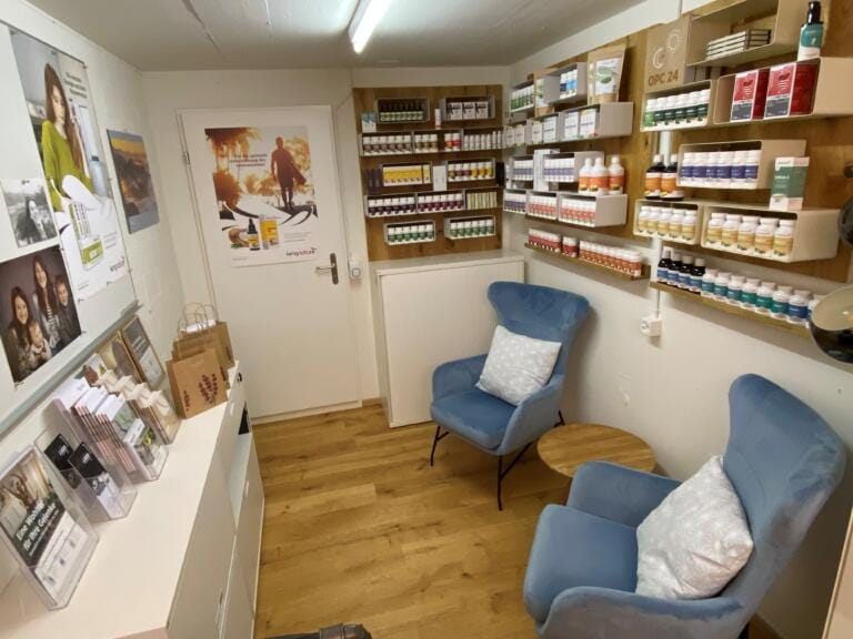 Ein gemütlicher Raum im Vitalshop Ost mit Holzboden, zwei blauen Sesseln mit weißen Kissen, einem kleinen runden Tisch und Regalen voller Vitamine und Nahrungsergänzungsmittel. Poster und Produktdisplays schmücken die Wände und die Theke.