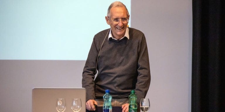 Ein älterer Mann mit Brille und Headset-Mikrofon steht lächelnd an einem Tisch mit Wasserflaschen und hält vor einem weißen Bildschirm eine Präsentation zum Thema Registrierung.